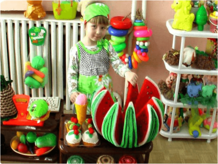 Девочка в зелёном костюме стоит около муляжа арбуза и другой еды