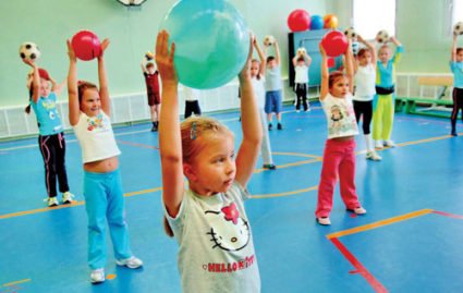 Дети в спортзале с синим полом держат мячи над головами